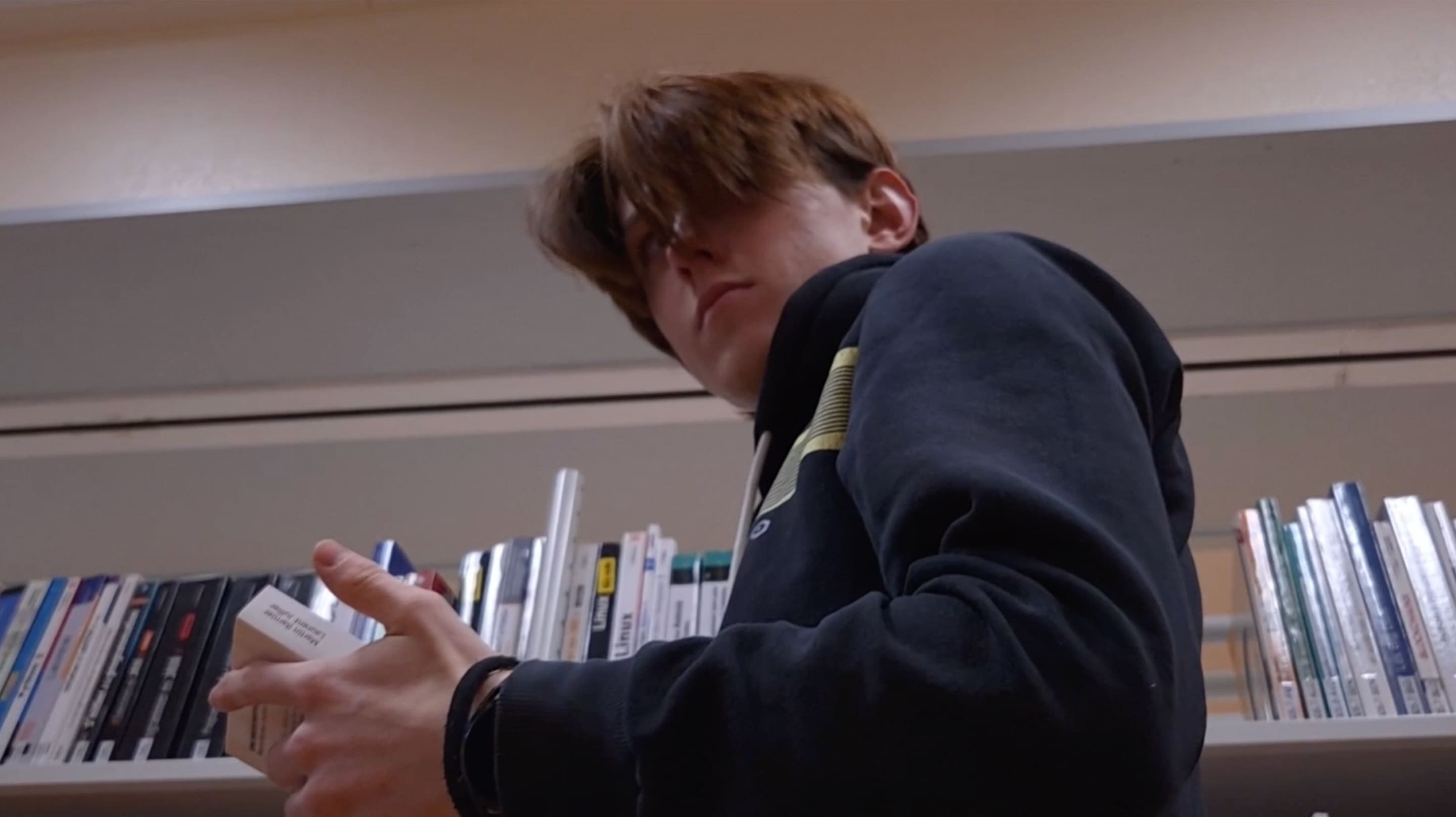 2e photographie de Presence où un homme tient un livre et regarde derrière lui d'un regard menaçant. Le plan est filmé en contre-plongée et se déroule dans une bibliothèque.