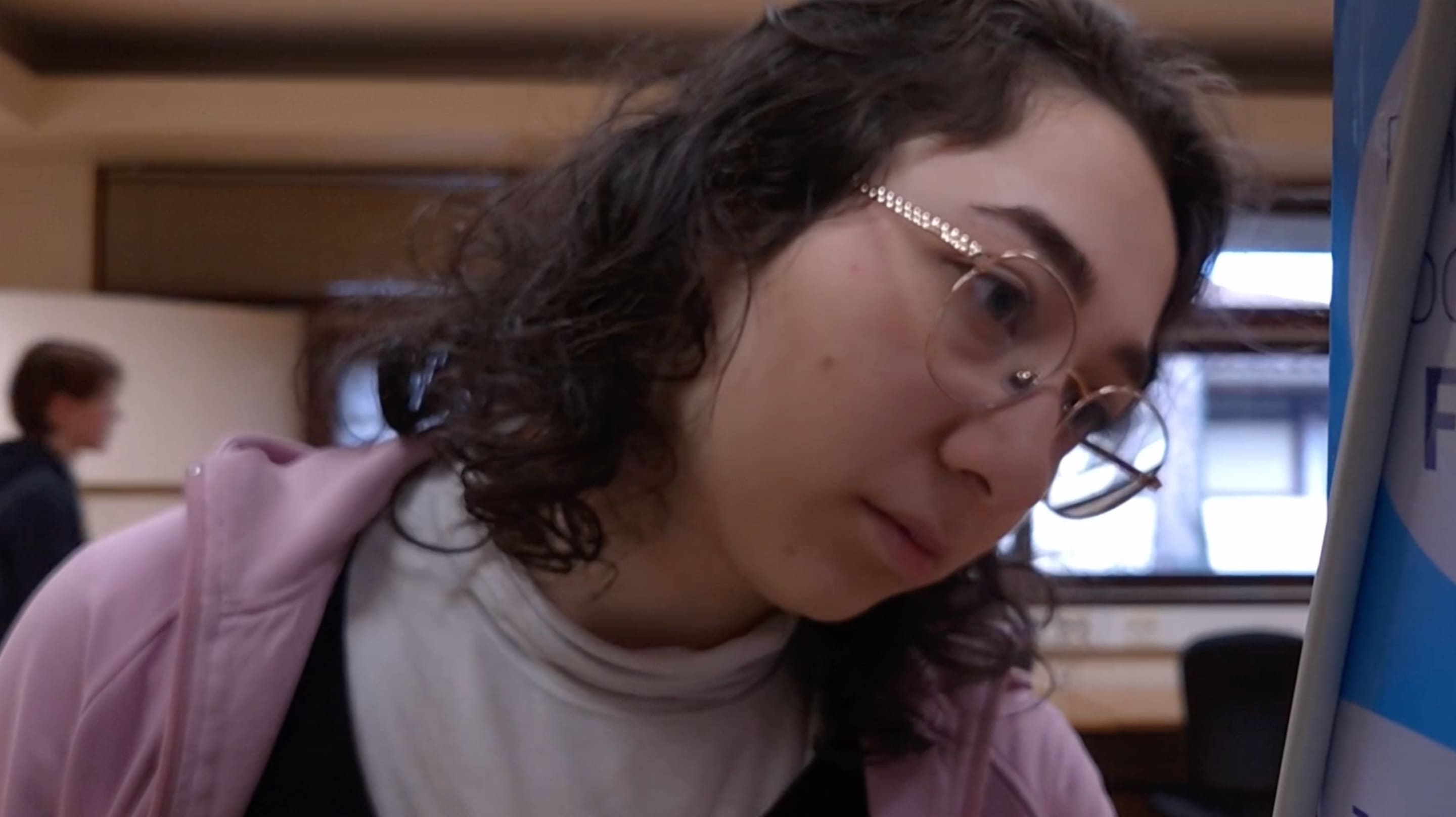 3e photographie de Presence où une femme regarde l'étagère d'une bibliothèque. Le plan est filmé de face et un homme se déplace derrière elle.